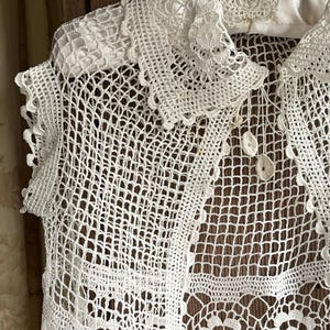May include: A white, open-weave crochet top with a detailed collar and floral accents. The intricate design features a delicate, lacy pattern, showcasing the craftsmanship of the handmade garment. The top is displayed on a hanger.
