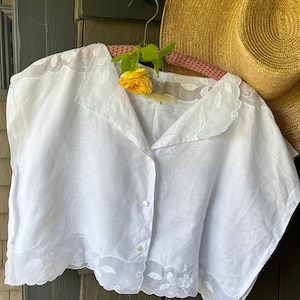 May include: A white, button-down blouse with delicate embroidered floral and leaf details along the collar and hem. A yellow rose and straw hat are in the background. The blouse is hanging on a pink hanger.