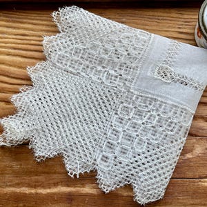 Puede incluir: Un pañuelo de encaje blanco con un intrincado diseño geométrico y un borde festoneado. La delicada tela está doblada sobre una superficie de madera. Un recipiente decorativo floral es visible en el fondo.