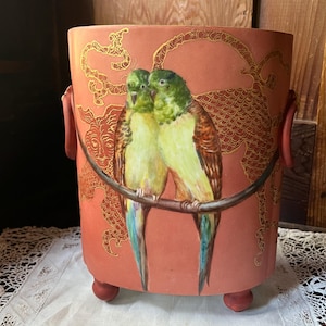 May include: A cylindrical terracotta-colored ceramic planter featuring two green parrots perched on a branch. The exterior is decorated with gold-toned line art patterns, two side handles, and three small rounded feet at the base.