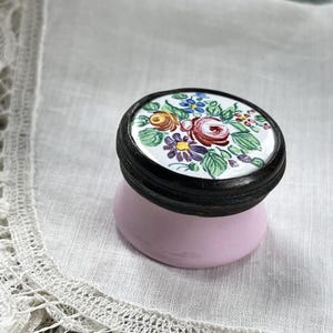 Op de afbeelding: Een kleine, antieke sieradendoos met een bloemmotief op het deksel. Het deksel heeft een witte achtergrond met een kleurrijk arrangement van bloemen, waaronder rode rozen, gele en blauwe bloemen. De basis is zachtroze.