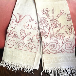 May include: Two off-white rectangular towels featuring red embroidered floral patterns. One towel displays poppy-like flowers above a geometric diamond border, while the other shows carnation-like blooms above a swirling vine design. Both have fringed edges.