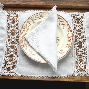 May include: A white linen placemat with intricate lace detailing and a matching napkin. A vintage plate with a floral design is placed on the placemat. The placemat and napkin are on a wooden surface, creating a rustic, elegant table setting.