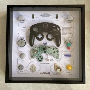 May include: A framed display of a disassembled Nintendo GameCube controller. The controller is black and features a white Nintendo logo. The display shows the individual components of the controller, including the buttons, joystick, and circuit board. The text "Nintendo GameCube" is printed on the display.
