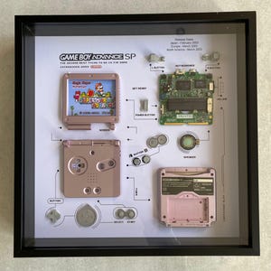 May include: A disassembled Game Boy Advance SP in a shadow box frame. The frame includes the motherboard, buttons, screen, and cartridge slot. The game cartridge is a Super Mario Advance 4 game.