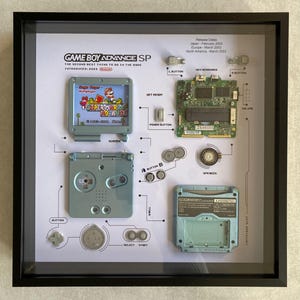 May include: A framed display of a disassembled Game Boy Advance SP, showing the individual components of the handheld gaming device. The display includes the motherboard, screen, buttons, and cartridge slot. The text on the display reads "Game Boy Advance SP, The Second Best Thing To Do In The Dark, Introduced 2003".