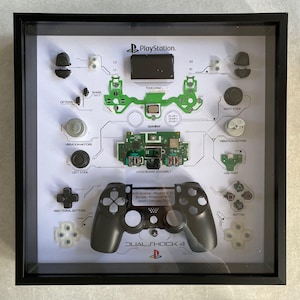 May include: A framed display of a disassembled PlayStation 4 DualShock 4 controller. The display includes the controller's individual components, such as the buttons, joysticks, and circuit board, arranged on a white background with a diagram of the controller's layout. The text "PlayStation" and "DualShock 4" are visible in the display.