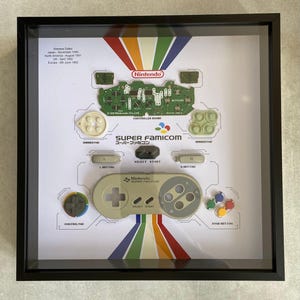 May include: A framed display of a disassembled Super Nintendo controller. The display includes the controller board, buttons, and a printed diagram of the controller layout. The text on the display reads "Super Famicom" and "Nintendo Super Famicom".