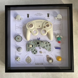 May include: A disassembled Nintendo GameCube controller displayed in a black frame. The controller is separated into its individual components, including the buttons, joystick, and circuit board. The text "Nintendo GameCube" is visible at the bottom of the frame.