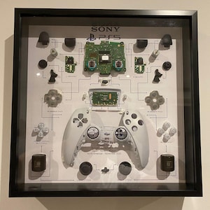 May include: A framed display of a deconstructed PlayStation 5 controller, revealing its internal parts. The arrangement includes the controller's shell, circuit boards, buttons, and joysticks, connected by lines. The Sony PS5 logo is at the top, with text indicating the region and date.