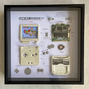 May include: A framed display of a disassembled Game Boy Advance SP, showing the individual components of the handheld gaming console. The display includes the motherboard, buttons, screen, and speaker. The text "GAME BOY ADVANCE SP" is printed at the top of the display.