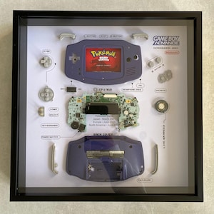 May include: A framed display of a disassembled Game Boy Advance. The purple handheld console is shown with its components, including the motherboard, buttons, and back cover. The screen displays "Pokémon Ruby Version." Text includes "Introduced 2001" and release dates.