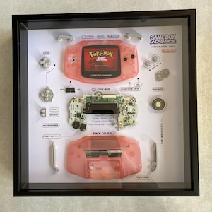 May include: A disassembled Game Boy Advance SP in a shadow box frame. The frame includes the console's shell, motherboard, buttons, and other components. The console is pink and the frame is black. The text "GAME BOY ADVANCE INTRODUCES 2001 Nintendo" is printed on the frame.