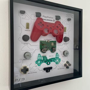 May include: A framed display of a disassembled PlayStation 3 DualShock 3 controller. The controller is shown in its component parts, including the buttons, joysticks, and circuit board. The display is labelled with the names of each part.