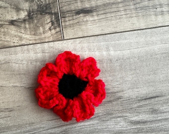 May include: A handcrafted, red crochet flower with a black centre. The flower features multiple petals and is set against a grey wooden background. This item could be used as an accessory or decoration.