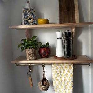 May include: Two light wood wall shelves with a metal rod underneath. The top shelf holds a patterned ceramic bottle, a yellow bowl, and a wooden cutting board. The bottom shelf holds two potted plants, a white pepper mill, and a glass oil cruet.