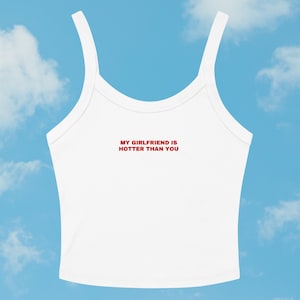 May include: White crop top with thin straps and the red text "MY GIRLFRIEND IS HOTTER THAN YOU". The top is made of a soft material and is designed for a comfortable fit. The background is a blue sky with white clouds.