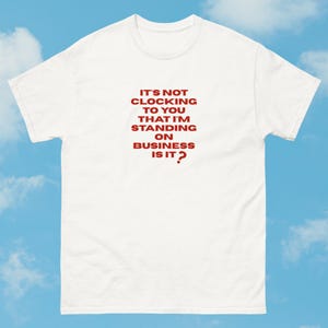 Könnte beinhalten: Weißes T-Shirt mit rotem Text: "IT'S NOT CLOCKING TO YOU THAT I'M STANDING ON BUSINESS IS IT?" Das Shirt hat einen Rundhalsausschnitt und kurze Ärmel. Der Hintergrund ist ein blauer Himmel mit weißen Wolken.