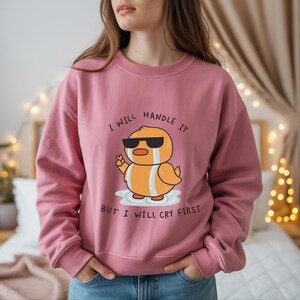 Puede incluir: Sudadera rosa polvorienta con un pato de dibujos animados con gafas de sol y llorando. El pato es naranja y blanco. El texto en la sudadera dice: "I WILL HANDLE IT BUT I WILL CRY FIRST".