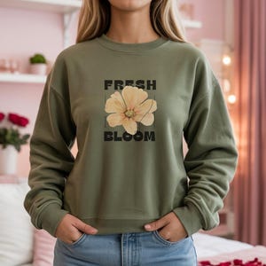 Pode incluir: Camisola verde-oliva com o texto "FRESH BLOOM" e um gráfico de flores bege. A camisola de gola redonda é de estilo casual e é feita de um material macio.