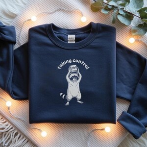 May include: Navy blue sweatshirt featuring a raccoon holding a "ctrl" button, with the words "taking control" above. The raccoon is in a standing position. The sweatshirt is on a white and beige surface with string lights.