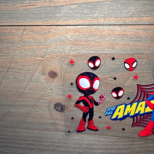 May include: A decorative cake topper featuring a cartoon Miles Morales character in a black and red suit, alongside the text "and his AMAZING" in yellow and blue, accented with small red and black stars and circular spider-mask icons.