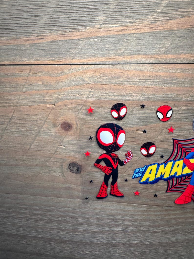 May include: A decorative cake topper featuring a cartoon Miles Morales character in a black and red suit, alongside the text "and his AMAZING" in yellow and blue, accented with small red and black stars and circular spider-mask icons.