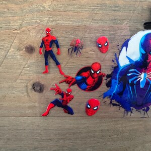 May include: A collection of Spider-Man themed stickers and cutouts. The stickers feature the superhero in various action poses, along with his mask and a spider emblem. The color scheme is primarily red, blue, and black, with a splash of blue paint.