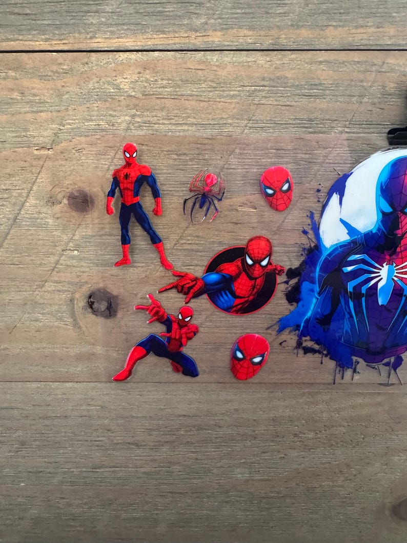 May include: A collection of Spider-Man themed stickers and cutouts. The stickers feature the superhero in various action poses, along with his mask and a spider emblem. The color scheme is primarily red, blue, and black, with a splash of blue paint.