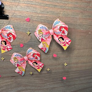 May include: Three hair bows featuring a pink fabric pattern with illustrations of various animated princesses, including characters with blonde, red, and black hair, accented with blue leaf motifs and small pink heart and gold star confetti.