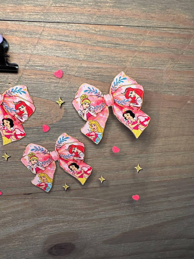 May include: Three hair bows featuring a pink fabric pattern with illustrations of various animated princesses, including characters with blonde, red, and black hair, accented with blue leaf motifs and small pink heart and gold star confetti.