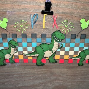 Puede incluir: Un recorte decorativo de papel que presenta al personaje de dinosaurio verde Rex de Toy Story, con el texto REX en letras coloridas. El dise&ntilde;o incluye un patr&oacute;n de cuadros en azul, turquesa y naranja, globos verdes y fuegos artificiales.