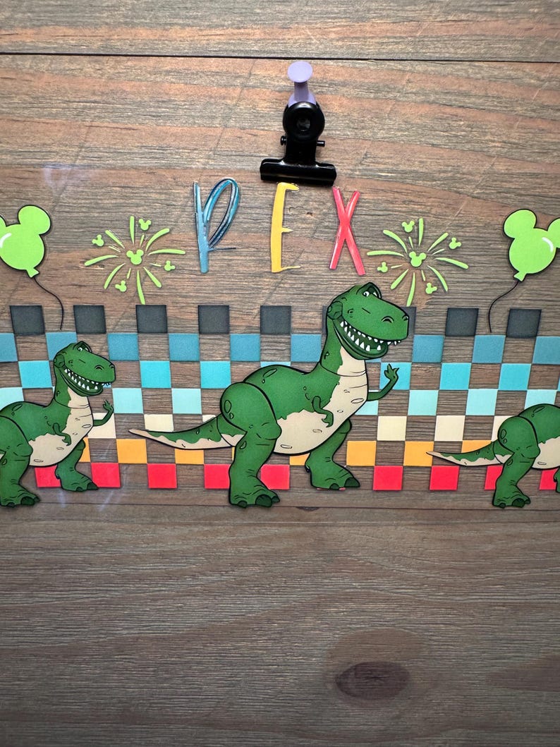 Puede incluir: Un recorte decorativo de papel que presenta al personaje de dinosaurio verde Rex de Toy Story, con el texto REX en letras coloridas. El dise&ntilde;o incluye un patr&oacute;n de cuadros en azul, turquesa y naranja, globos verdes y fuegos artificiales.