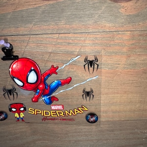 K&ouml;nnte beinhalten: Eine transparente Kunststofffolie mit einem Cartoon-Spider-Man in einem rot-blauen Anzug mit wei&szlig;en Spinnennetzlinien. Das Marvel Spider-Man Homecoming-Logo befindet sich darunter, zusammen mit Spinnen-Symbolen und einer kleinen Spider-Man-Figur.