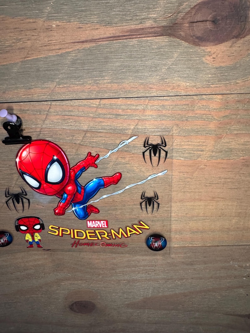 K&ouml;nnte beinhalten: Eine transparente Kunststofffolie mit einem Cartoon-Spider-Man in einem rot-blauen Anzug mit wei&szlig;en Spinnennetzlinien. Das Marvel Spider-Man Homecoming-Logo befindet sich darunter, zusammen mit Spinnen-Symbolen und einer kleinen Spider-Man-Figur.
