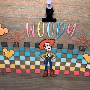 May include: A decorative sign featuring the character Woody from Toy Story, centered above a colorful checkered pattern. The word WOODY is printed in multi-colored letters above the character. The design includes orange Mickey-shaped balloons and fireworks.