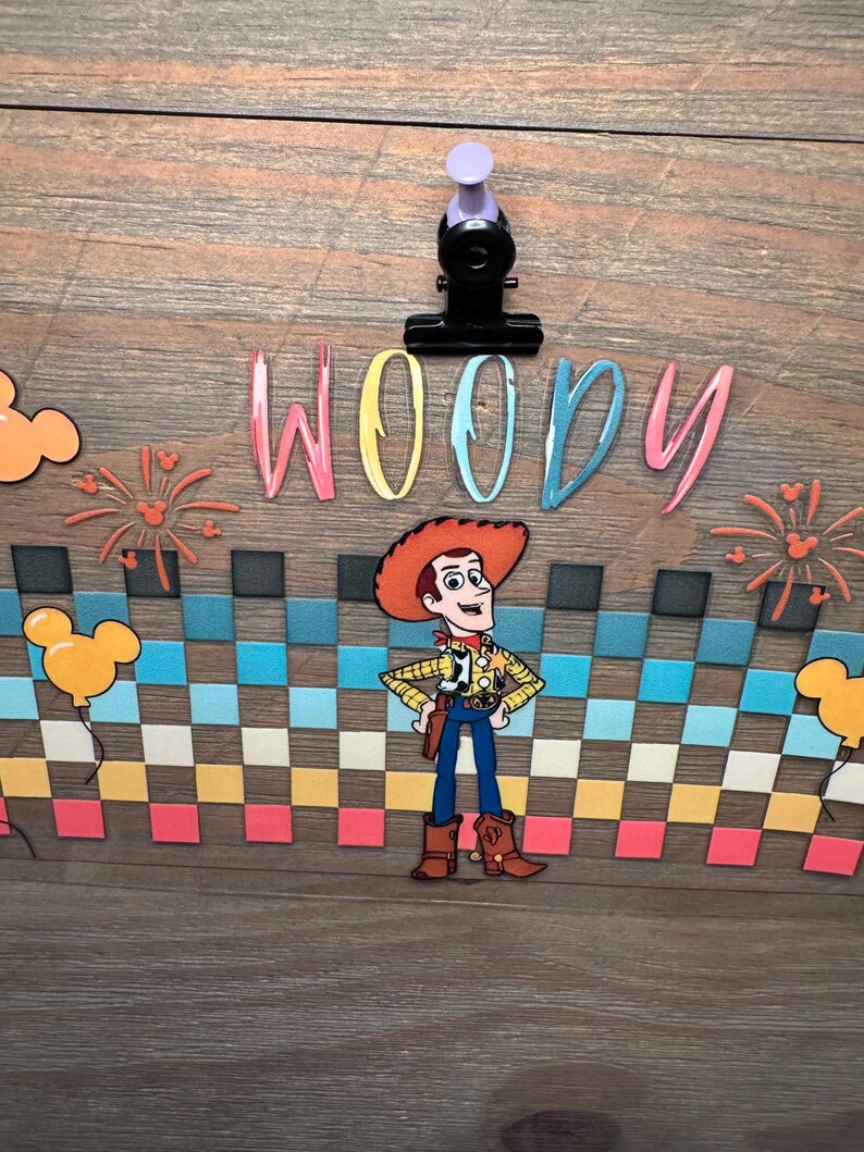 May include: A decorative sign featuring the character Woody from Toy Story, centered above a colorful checkered pattern. The word WOODY is printed in multi-colored letters above the character. The design includes orange Mickey-shaped balloons and fireworks.