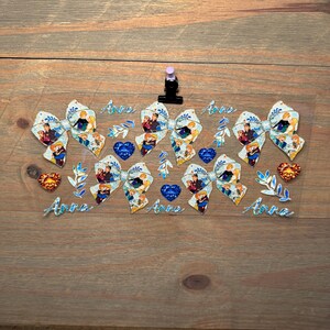 May include: A clear sheet featuring four glittery bows with a cartoon character print, four blue heart-shaped gems, two orange heart-shaped gems, several blue leaf sprigs, and the name Anna written in a blue cursive font four separate times.