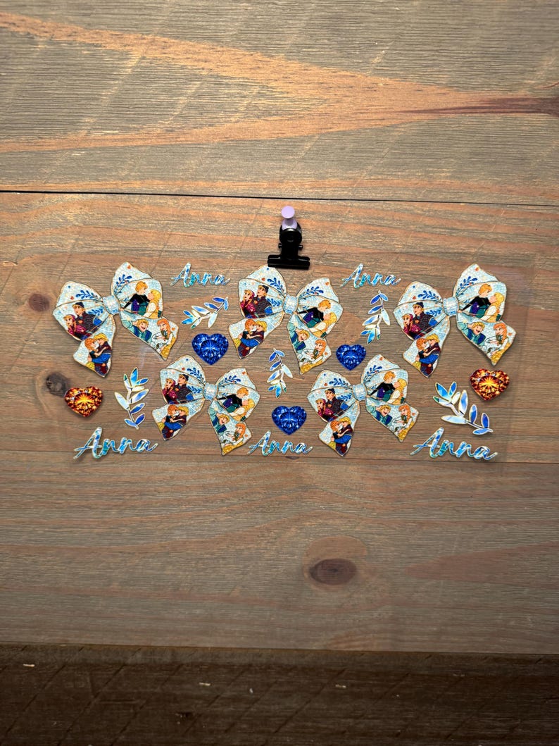 May include: A clear sheet featuring four glittery bows with a cartoon character print, four blue heart-shaped gems, two orange heart-shaped gems, several blue leaf sprigs, and the name Anna written in a blue cursive font four separate times.