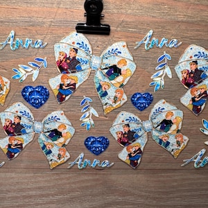 May include: A collection of light blue hair bows featuring a repeating pattern of cartoon characters, accompanied by blue heart-shaped gems, blue leaf-shaped accents, and the name Anna written in blue script, all secured by a black metal clip.