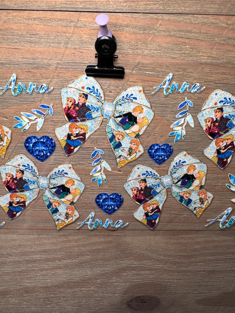 May include: A collection of light blue hair bows featuring a repeating pattern of cartoon characters, accompanied by blue heart-shaped gems, blue leaf-shaped accents, and the name Anna written in blue script, all secured by a black metal clip.