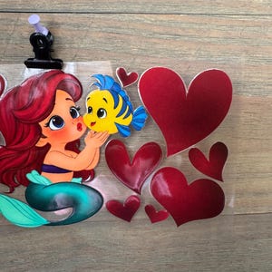 May include: Ariel and Flounder kiss sticker with red hearts. Ariel has red hair and a teal tail. Flounder is yellow and blue. The hearts are various sizes and shades of red. The sticker is held in place with a black clip.