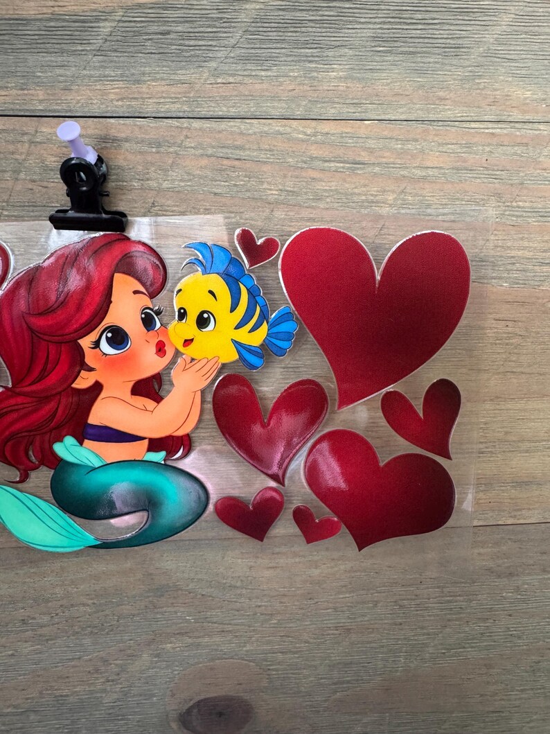 May include: Ariel and Flounder kiss sticker with red hearts. Ariel has red hair and a teal tail. Flounder is yellow and blue. The hearts are various sizes and shades of red. The sticker is held in place with a black clip.