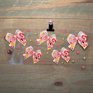 May include: Five fabric hair bows featuring a repeating pattern of various cartoon princesses in pink, yellow, and blue tones. The bows are arranged on a clear sheet decorated with small pink heart and gold star confetti accents.