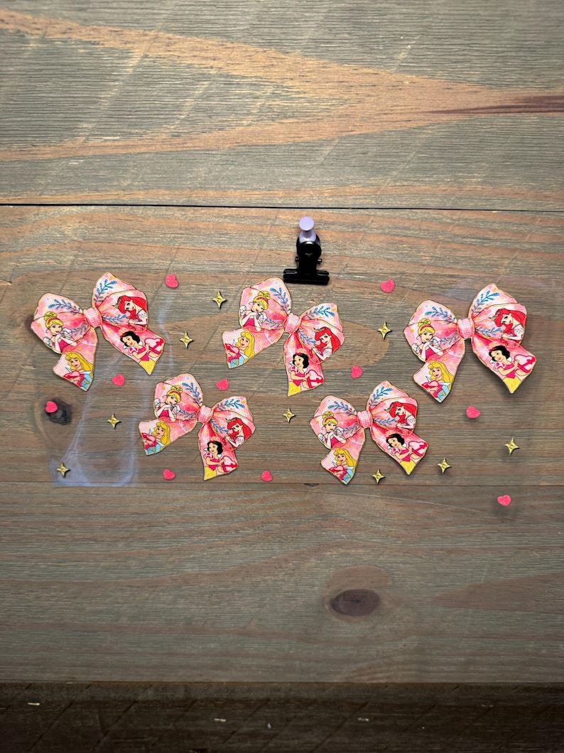 May include: Five fabric hair bows featuring a repeating pattern of various cartoon princesses in pink, yellow, and blue tones. The bows are arranged on a clear sheet decorated with small pink heart and gold star confetti accents.