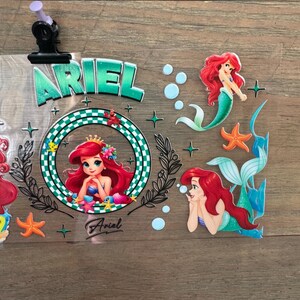 Pu&ograve; includere: Adesivi a tema Ariel con la Sirenetta. Gli adesivi includono il nome "Ariel" in lettere verdi, immagini di Ariel, stelle marine e bolle. Gli adesivi sono su uno sfondo trasparente.