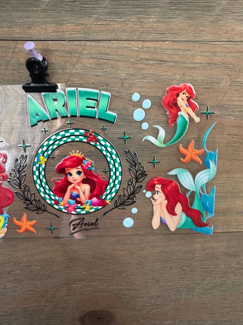 Pu&ograve; includere: Adesivi a tema Ariel con la Sirenetta. Gli adesivi includono il nome "Ariel" in lettere verdi, immagini di Ariel, stelle marine e bolle. Gli adesivi sono su uno sfondo trasparente.