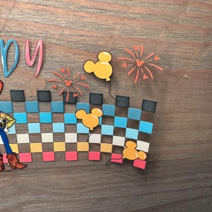 May include: A clear adhesive decal featuring a partial view of a character in a yellow shirt and blue pants, a checkered pattern in shades of blue, yellow, and pink, three yellow Mickey Mouse-shaped balloons, and orange firework illustrations.
