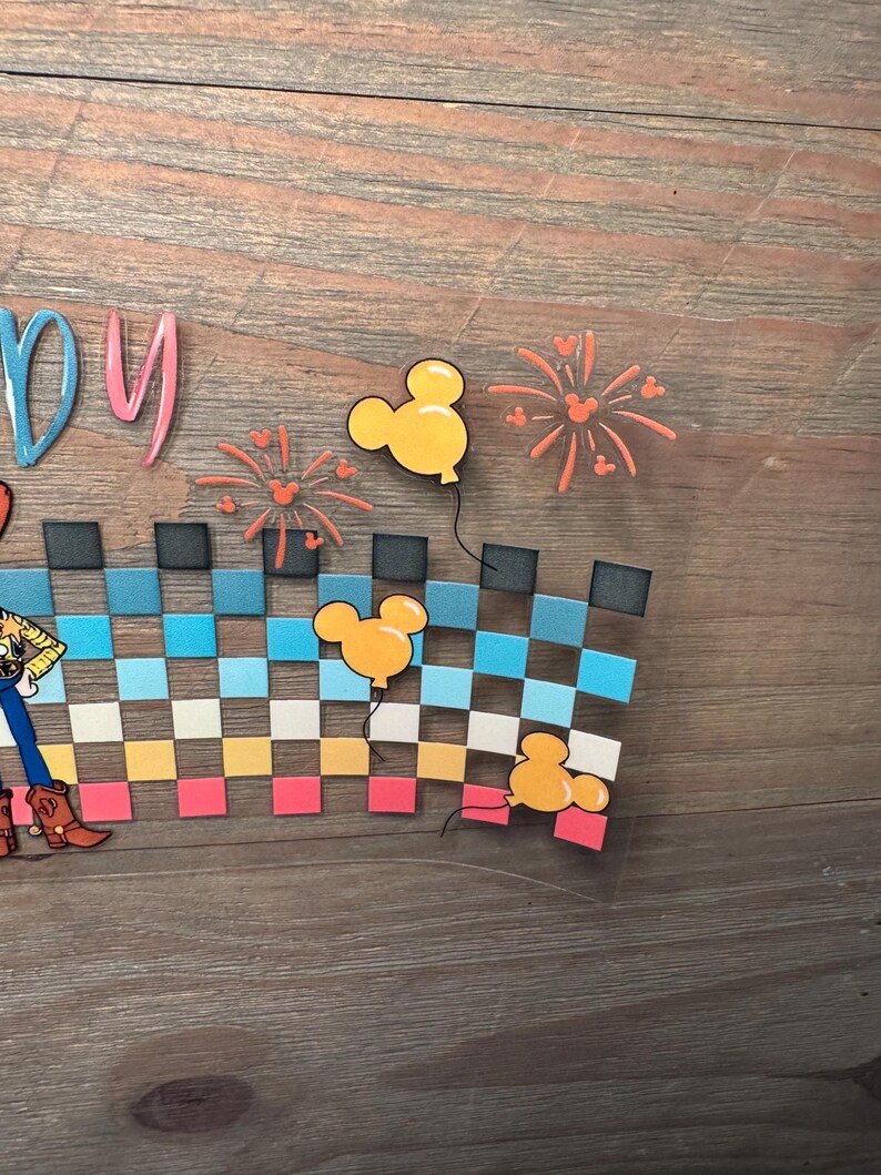 May include: A clear adhesive decal featuring a partial view of a character in a yellow shirt and blue pants, a checkered pattern in shades of blue, yellow, and pink, three yellow Mickey Mouse-shaped balloons, and orange firework illustrations.