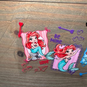 May include: Two die-cut stickers featuring a cartoon mermaid with red hair and a teal tail. One mermaid sits on a rock, the other waves. The stickers include pink striped backgrounds, red and purple hearts, arrows, a paper airplane, and the text Be MINE.